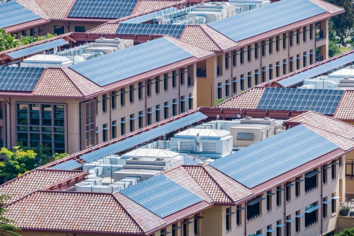 Rozległy kompleks budynków z licznymi panelami fotowoltaicznymi na dachach, zaspokajający potrzeby energetyczne dużej wspólnoty mieszkaniowej.