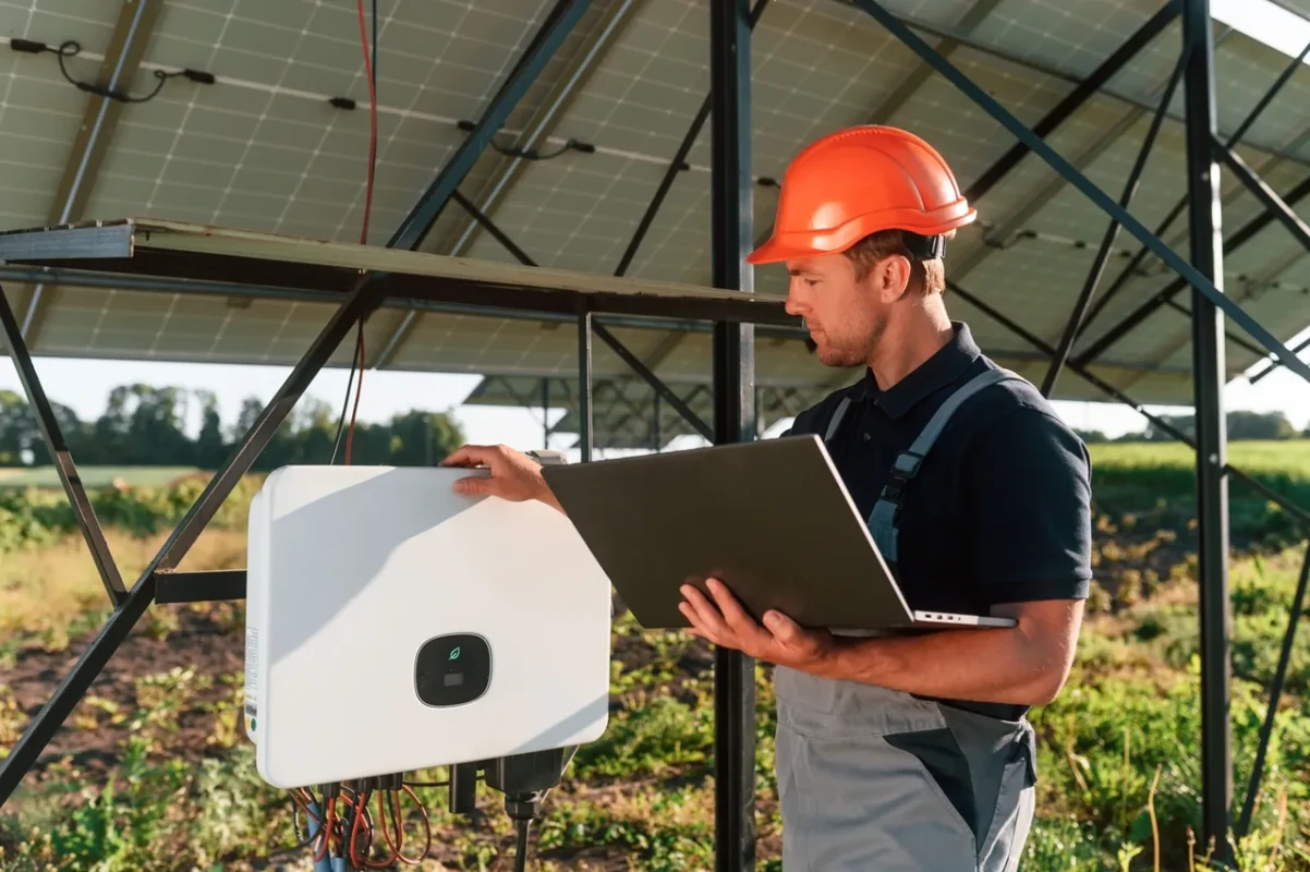 Technik w pomarańczowym kasku konfiguruje falownik fotowoltaiczny na laptopie, przygotowując go do podłączenia magazynu energii LV.