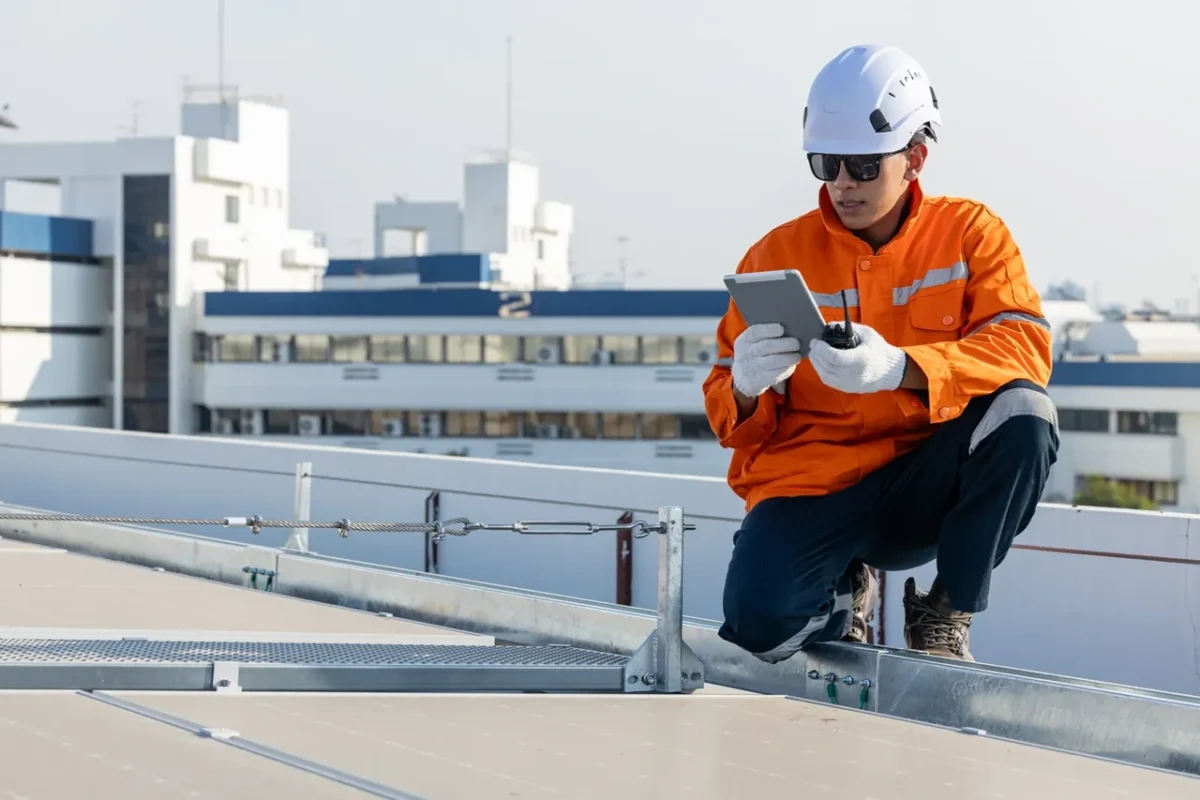 Technik w pomarańczowym stroju sprawdza konstrukcję montażową na dachu, przygotowując instalację PV dla sklepu i warsztatu.