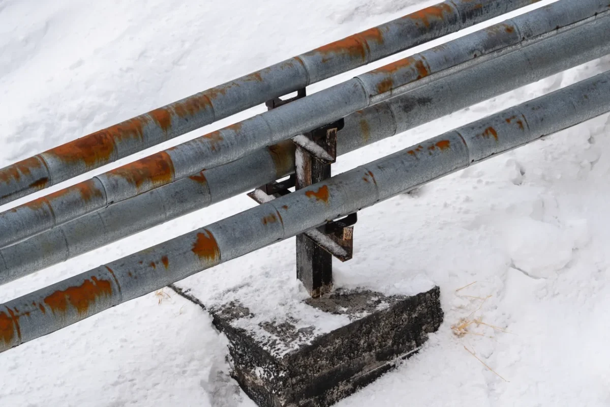 Ocynkowane rury stalowe z plamami rdzy zainstalowane na zewnątrz, w zaśnieżonym miejscu, pokazujące uszkodzenia korozyjne elementów metalowych w trudnych warunkach klimatycznych.