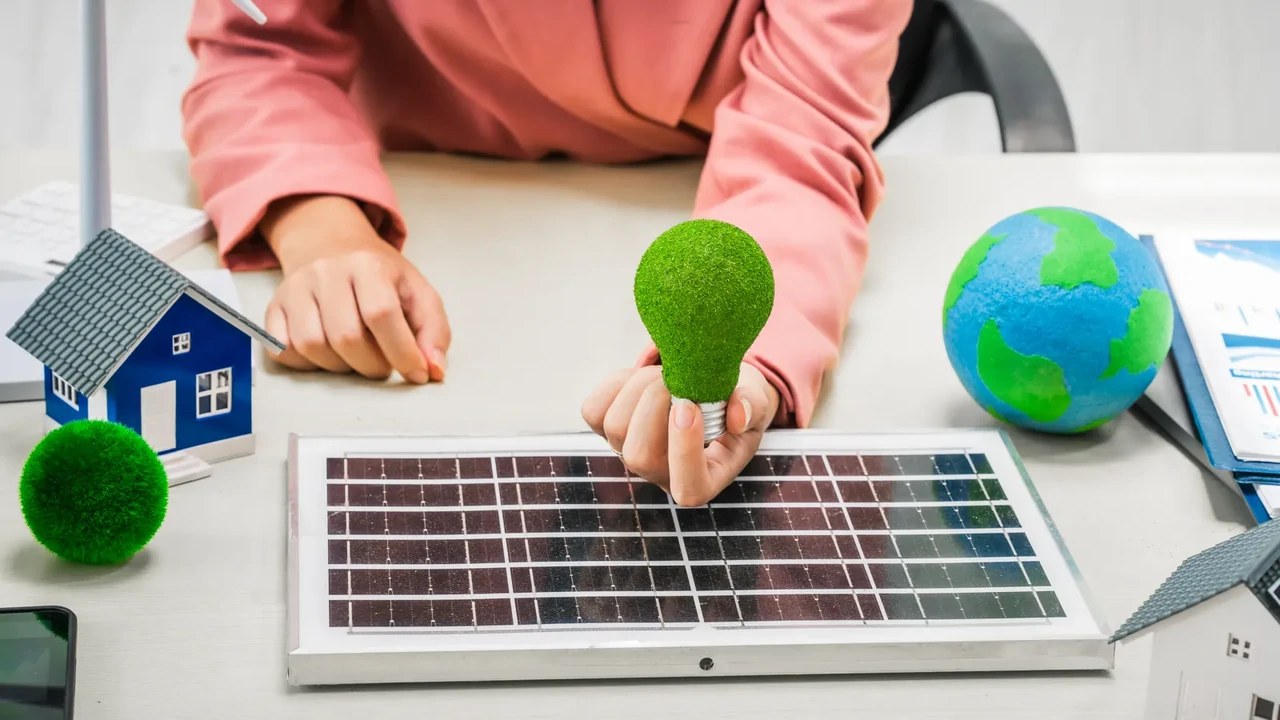 Ręka trzyma zieloną żarówkę nad panelem PV, ilustrując koncepcję deratingu inwertera jako utraty wydajności energetycznej.