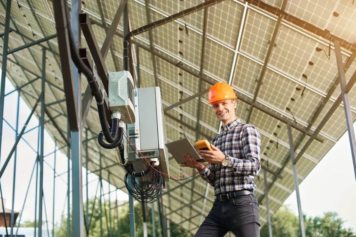 Uśmiechnięty inżynier trzymający tablet i tester obok falownika fotowoltaicznego, prezentujący profesjonalną obsługę i serwis posprzedażowy systemów energii słonecznej
