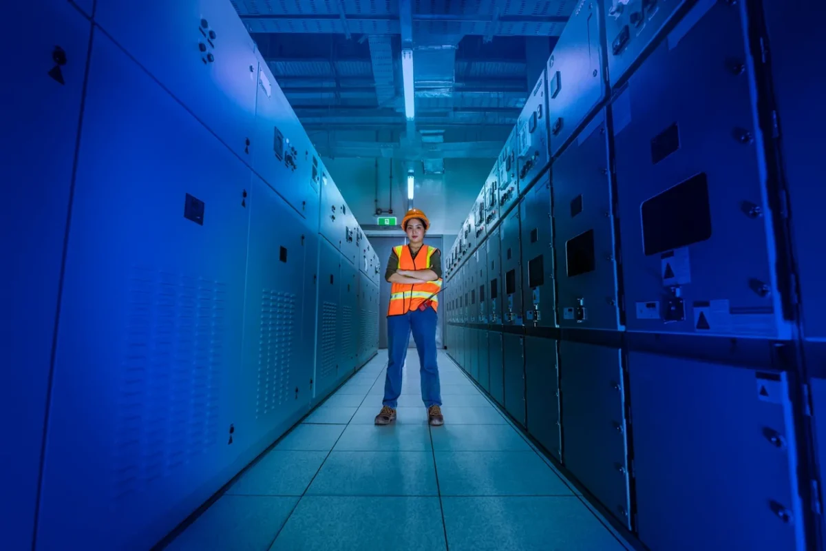Technik w pomarańczowym kasku w sali rozdzielczej, demonstrujący zgodność montażu magazynu energii z normami bezpieczeństwa.