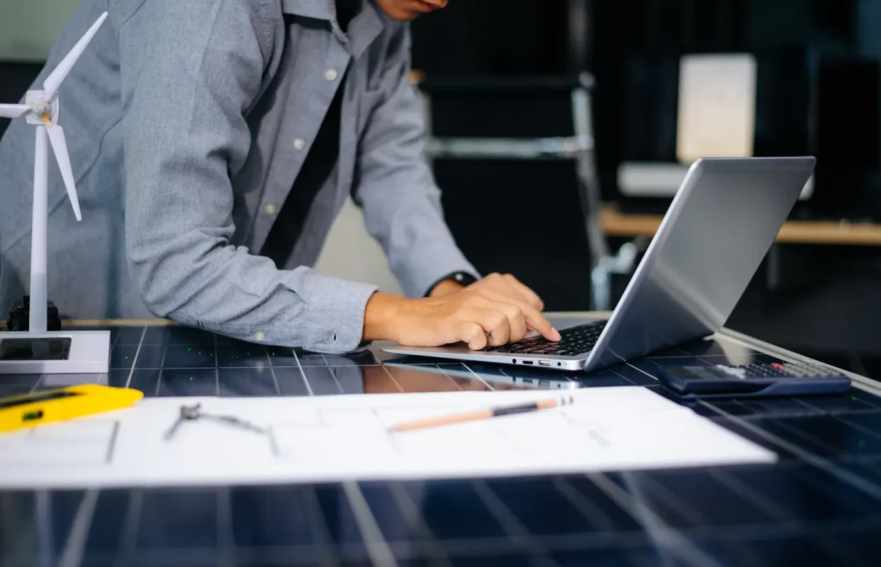 Inżynier projektuje firmową instalację fotowoltaiczną na laptopie, na stole leżą schematy, model turbiny wiatrowej oraz fragment panelu słonecznego.