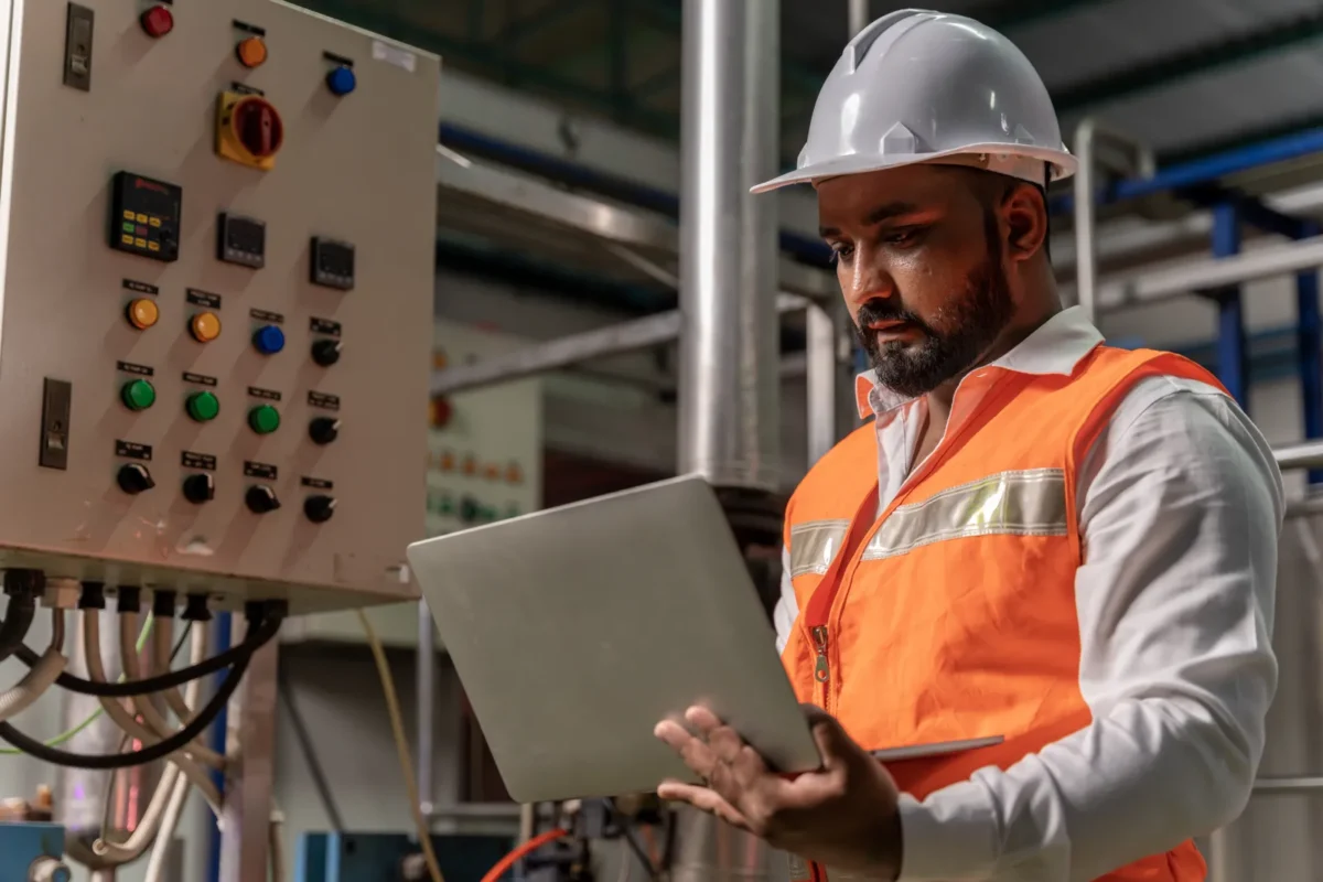 Technik w kamizelce ostrzegawczej sprawdza parametry instalacji elektrycznej na laptopie przy szafie sterującej, kontrolując pracę falowników i magazynów energii.