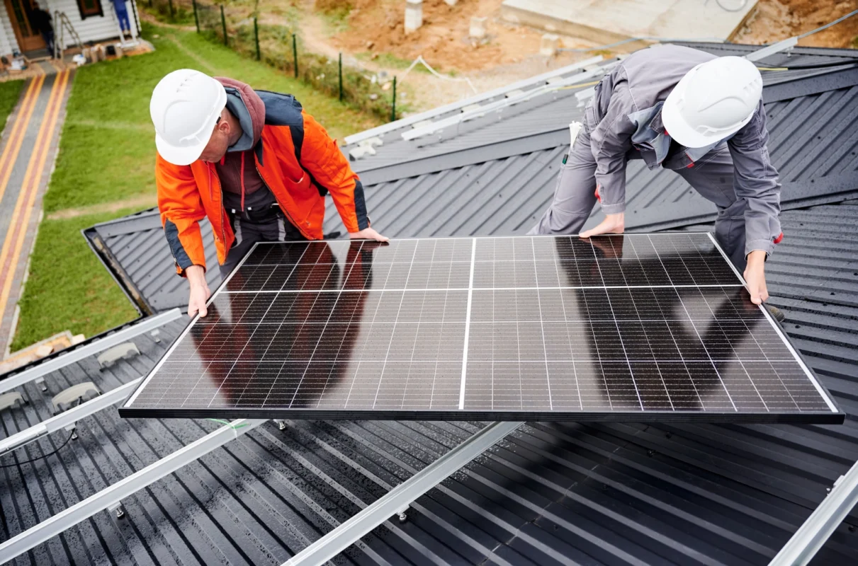 Dwóch instalatorów umieszcza duży panel fotowoltaiczny na blaszanym dachu podczas montażu domowej instalacji PV typu off-grid.