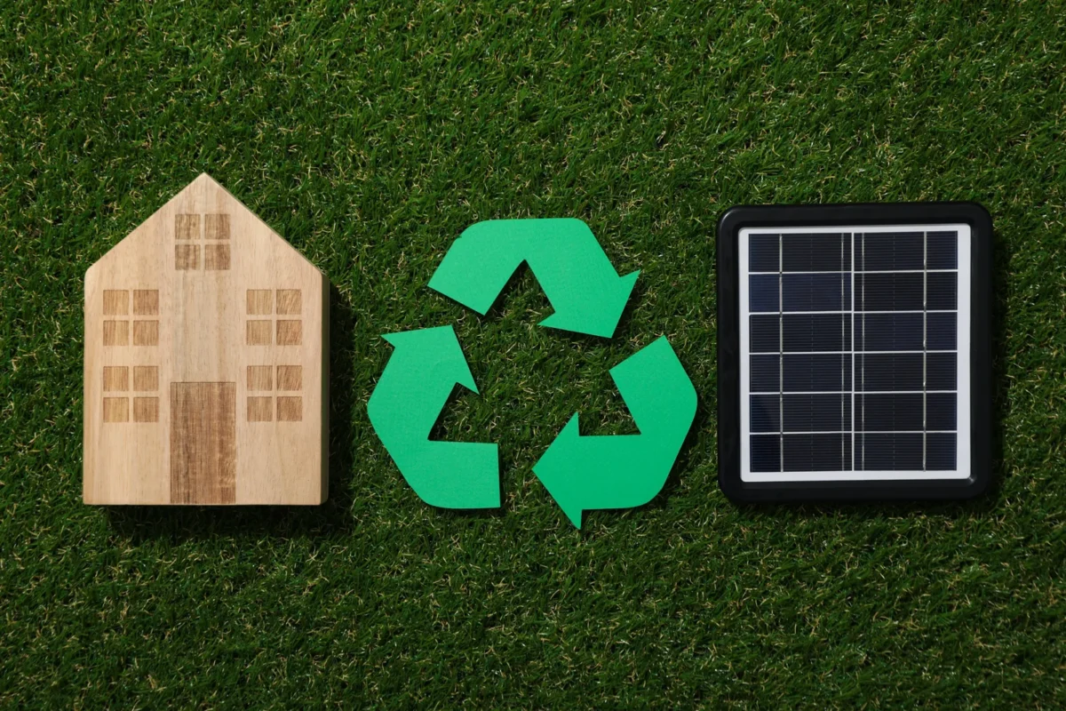 Drewniany model domu, znak recyklingu i mała bateria słoneczna ułożone na zielonej trawie, symbolizują gospodarkę nadwyżką energii z domowej instalacji fotowoltaicznej.