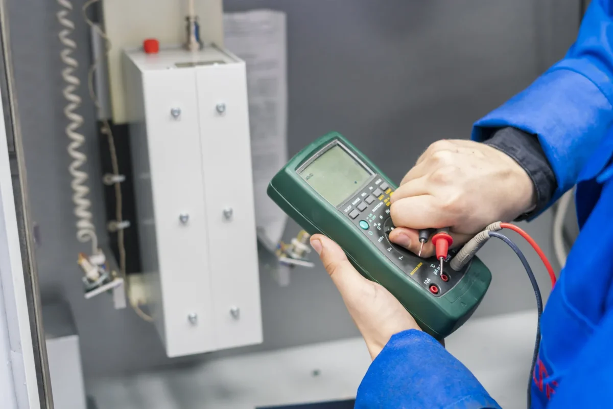 Elektryk w niebieskiej odzieży roboczej używa multimetru do pomiaru parametrów elektrycznych przed wykonaniem prac mających na celu obniżenie napięcia instalacji.